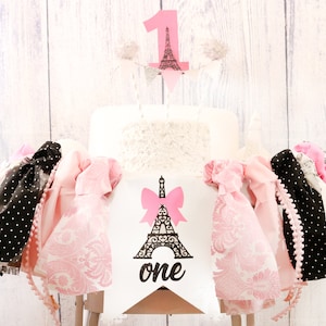 May include: A white cake with pink and black decorations, including a pink Eiffel Tower and a banner that says "one". The cake is surrounded by pink and black fabric and a white wooden chair.