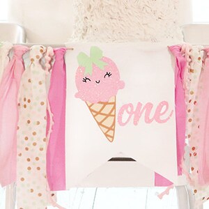 May include: A high chair banner with a pink ice cream cone graphic and the word "one". The banner is decorated with ribbons in pink, gold, and mint green. The ribbons are of varying textures, including polka dots and glitter.