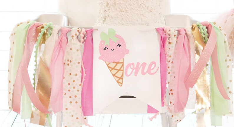 May include: A high chair banner with a pink ice cream cone graphic and the word "one". The banner is decorated with ribbons in pink, gold, and mint green. The ribbons are of varying textures, including polka dots and glitter.