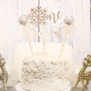 Può includere: Una torta bianca glassata con un topper decorativo che dice "one" e un fiocco di neve dorato glitterato. La torta è decorata con uno striscione e pompon. Due statuette di renne dorate e palline decorative affiancano la torta.