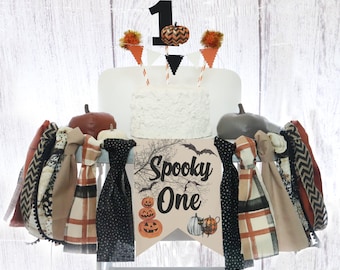 May include: A white cake with orange and black decorations for a first birthday party. The cake is decorated with a black number one, orange and black pumpkin decorations, and a banner that says "Spooky One".