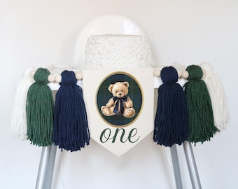 Puede incluir: Un banderín para silla alta con un estandarte central color crema que muestra una imagen circular de un oso de peluche con un lazo azul marino sobre un fondo de cuadros verde oscuro. La palabra one aparece debajo en letra cursiva.