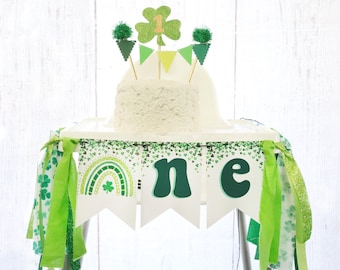 May include: Green and white high chair banner with shamrocks, a rainbow, and the word "one" in green letters. The banner is decorated with green and white ribbons.