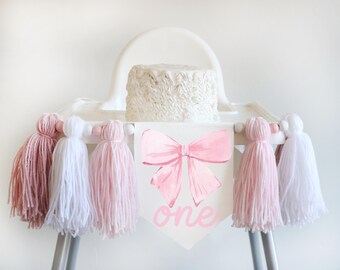 May include: A white high chair with a pink and white tassel garland. A white cake with pink frosting sits on the chair. A banner with a pink bow and the word "one" is attached to the chair.