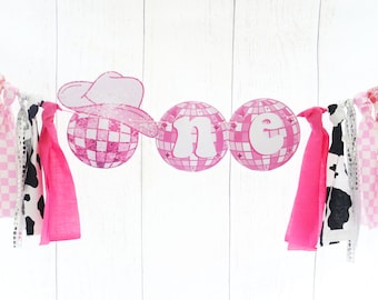 May include: Pink and white fabric banner with the word "one" in white letters on pink disco ball shapes. The banner has a pink cowboy hat and pink and white fabric streamers.
