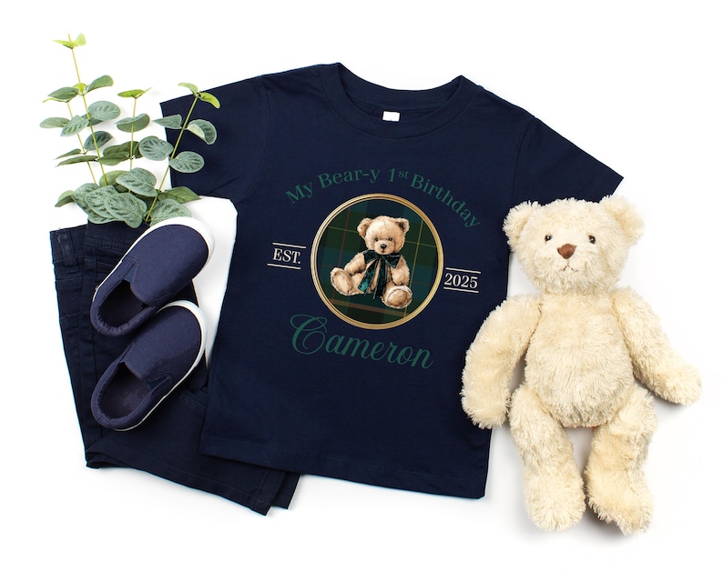 May include: Navy blue t-shirt with a teddy bear graphic and the text "My Bear-y 1st Birthday Cameron 2025". The image also includes a pair of navy blue slip-on shoes, navy blue pants, and a stuffed teddy bear.