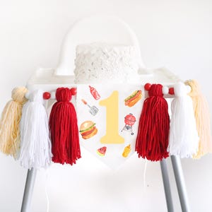 May include: A white high chair banner featuring a large yellow number one surrounded by watercolor illustrations of a burger, hot dog, grill, spatula, ketchup, mustard, and watermelon, accented with red, white, and yellow yarn tassels.