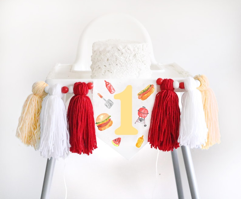May include: A white high chair banner featuring a large yellow number one surrounded by watercolor illustrations of a burger, hot dog, grill, spatula, ketchup, mustard, and watermelon, accented with red, white, and yellow yarn tassels.