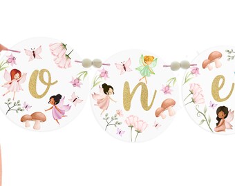May include: A white and gold "one" banner with a fairy theme. The banner is made of round circles with a gold glitter letter and fairy illustrations. The fairies are wearing pink, green, and blue dresses with wings. The banner is decorated with pink flowers, butterflies, and mushrooms.
