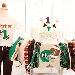 May include: A white onesie with brown and green football-themed graphics and the text "Parker is #1". A white cake with a brown number one topper and green and brown football-themed decorations. A brown and green fabric garland with football-themed graphics.