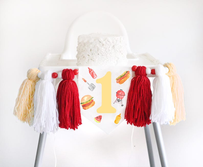 May include: A white high chair banner featuring a large yellow number one surrounded by watercolor illustrations of a burger, hot dog, grill, spatula, ketchup, mustard, and watermelon, accented with red, white, and yellow yarn tassels.