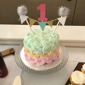 May include: A tiered birthday cake with pastel frosting in shades of pink, yellow, and mint green. The cake is decorated with a pink number one cake topper, gold and teal pennant banner, and silver pom-pom accents. It sits on a glass cake stand.