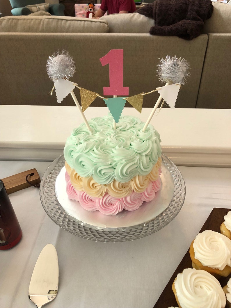 May include: A tiered birthday cake with pastel frosting in shades of pink, yellow, and mint green. The cake is decorated with a pink number one cake topper, gold and teal pennant banner, and silver pom-pom accents. It sits on a glass cake stand.