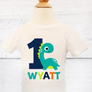 Pode incluir: Camiseta branca de manga curta com um n&uacute;mero um azul e um dinossauro verde. O dinossauro tem espinhos amarelos e p&eacute;s laranja. O texto "WYATT" est&aacute; impresso abaixo do dinossauro em verde, azul e laranja.