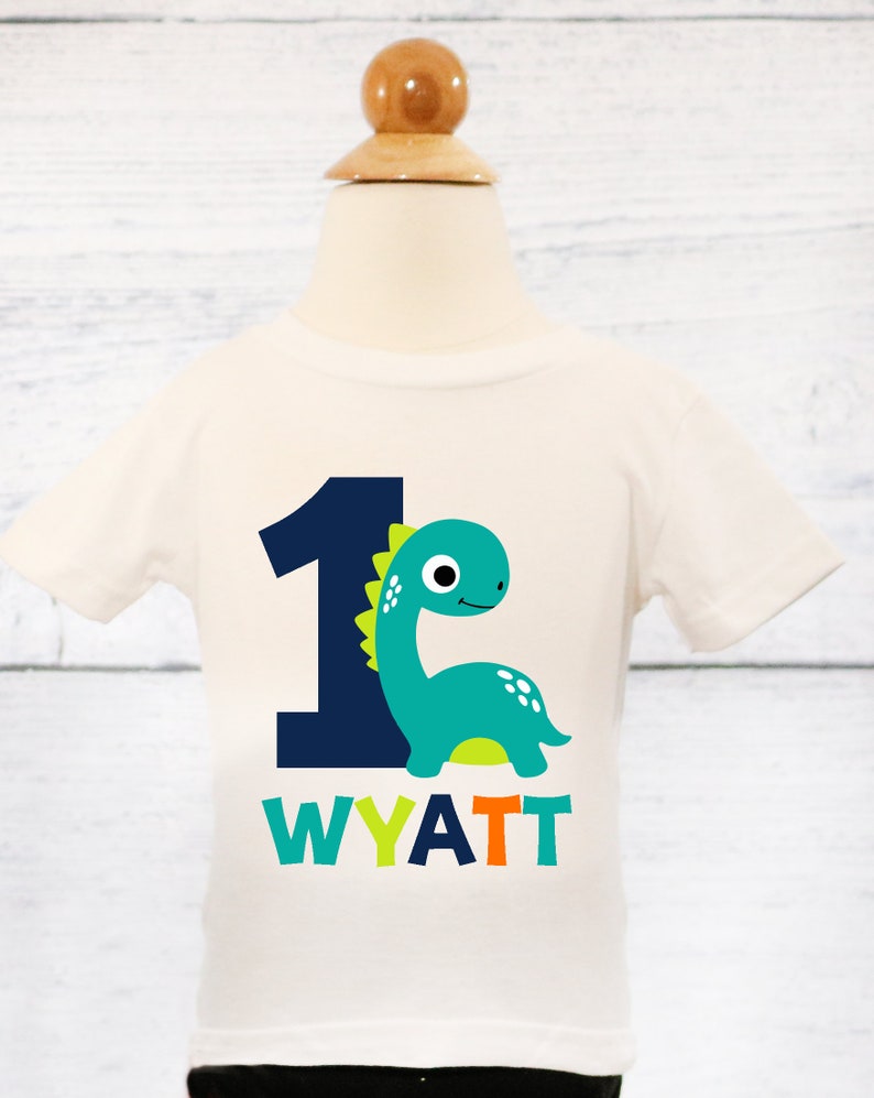 Pode incluir: Camiseta branca de manga curta com um n&uacute;mero um azul e um dinossauro verde. O dinossauro tem espinhos amarelos e p&eacute;s laranja. O texto "WYATT" est&aacute; impresso abaixo do dinossauro em verde, azul e laranja.