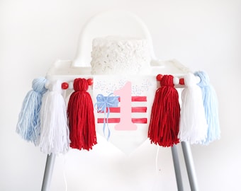 May include: A white high chair with a red, white, and blue banner that says "1" and has a watercolor design. The banner is decorated with red, white, and blue yarn tassels.