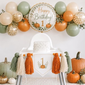 May include: A high chair decorated with a white banner featuring a pumpkin and the number 1, accented by orange, cream, and green yarn tassels. Above, a balloon arch includes sage green, cream, orange, and gold confetti balloons with a central balloon.