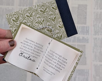 May include: A small, open book with printed text, held against a background of printed pages. The book has a patterned cover in olive green and white, with a navy blue spine. The text inside is about Christmas.