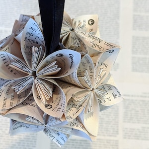 May include: A cluster of paper flowers made from the pages of an old book. The flowers are arranged in a sphere shape and are suspended from a black string.