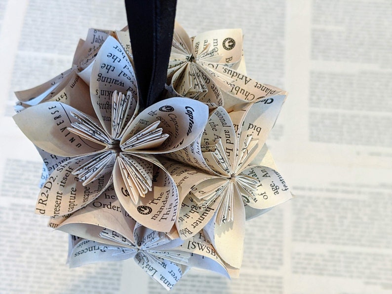 May include: A cluster of paper flowers made from the pages of an old book. The flowers are arranged in a sphere shape and are suspended from a black string.