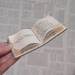 May include: A miniature open book titled Pride & Prejudice by Jane Austen with cream-colored pages and black printed text. The book is held open by a thin black and white twine string. The visible text includes the famous confession of love.