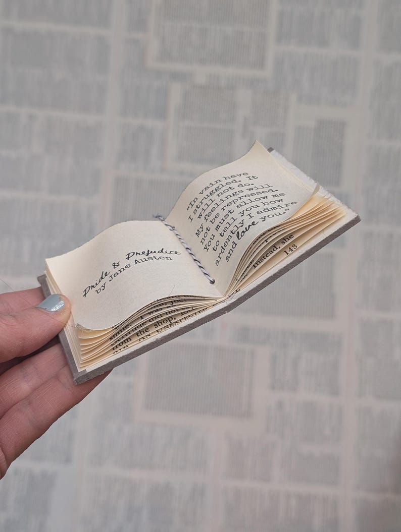 May include: A miniature open book titled Pride & Prejudice by Jane Austen with cream-colored pages and black printed text. The book is held open by a thin black and white twine string. The visible text includes the famous confession of love.