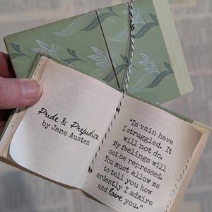 May include: A miniature book titled Pride & Prejudice by Jane Austen with a sage green patterned cover. The open pages display the text: In vain have I struggled. It will not do. My feelings will not be repressed. You must allow me to tell you how ardently I admire and love you. The book is bound with black and white twine.