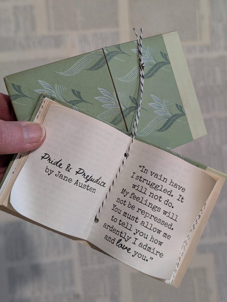 May include: A miniature book titled Pride & Prejudice by Jane Austen with a sage green patterned cover. The open pages display the text: In vain have I struggled. It will not do. My feelings will not be repressed. You must allow me to tell you how ardently I admire and love you. The book is bound with black and white twine.