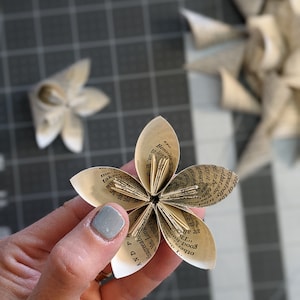 May include: A paper flower made from the pages of an old book. The flower has six petals and is held in a hand with a light blue nail polish.