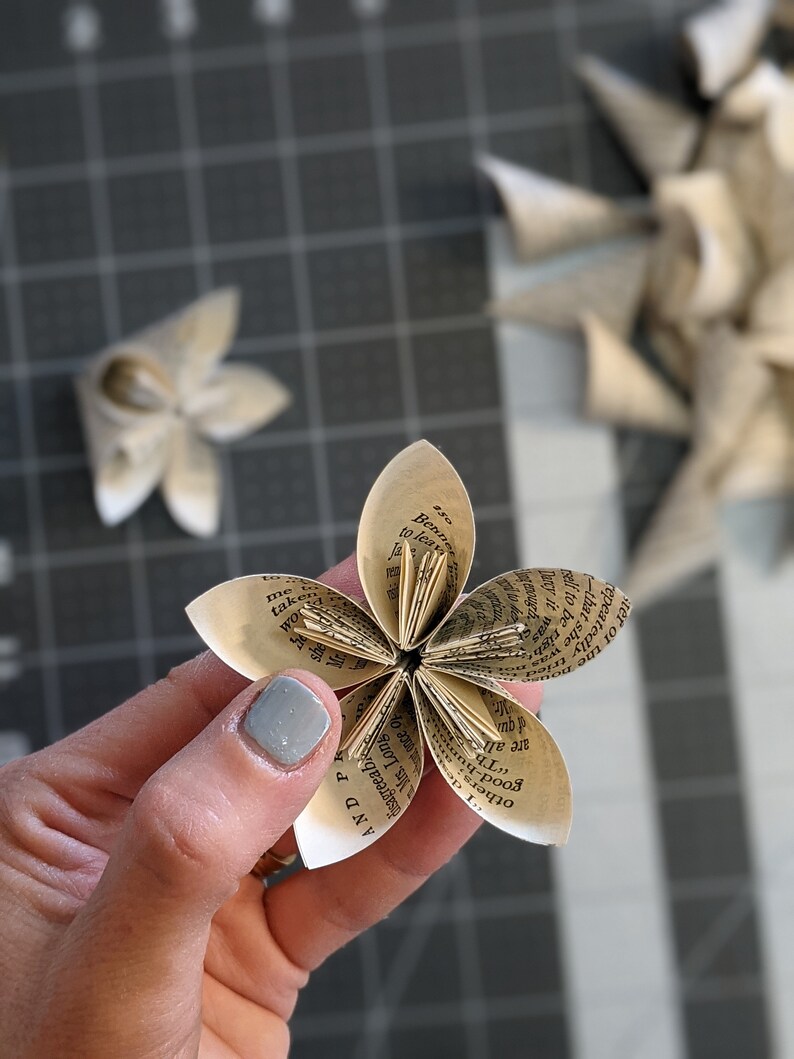 May include: A paper flower made from the pages of an old book. The flower has six petals and is held in a hand with a light blue nail polish.