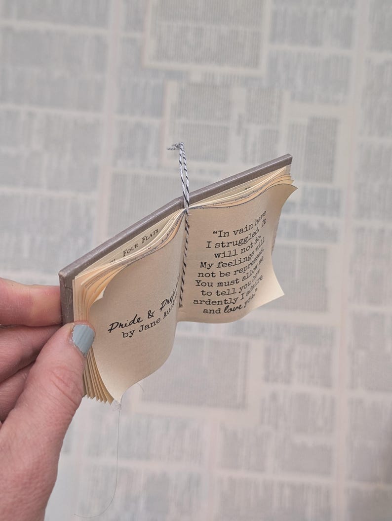 May include: A miniature book titled Pride & Prejudice by Jane Austen with a grey cover and black and white twine binding. The open pages display the text: In vain have I struggled. It will not do. My feelings will not be repressed.