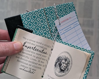 May include: A miniature book with a teal and white geometric patterned cover, tied with black and white twine. The open pages feature printed text about a lion and an illustration of a lion, with a library checkout card labeled bookology co.