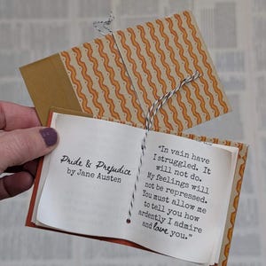 May include: A miniature book titled Pride & Prejudice by Jane Austen with a patterned orange and white wavy cover. The open pages display the text: In vain have I struggled. It will not do. My feelings will not be repressed. You must allow me to tell you how ardently I admire and love you. The book is bound with black and white twine.