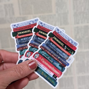 May include: Three stackable stickers featuring a stack of books with Christmas-themed titles. The stickers are white with a blue and red colour scheme. The titles include "A Christmas Carol", "Christmas with Anne", "Emmanuel", "The Best Christmas Stories Ever", "Dickens of Christmas", "Charles Dickens", "Shadow & Light", "A Child's Christmas in Wales", "Christmas Album", "A Christmas Treasury", "Twas The Night Before Christmas", "By Words on a Snowy Evening", and "The Real Book Snowman".