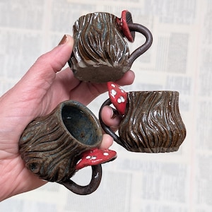 May include: Three small ceramic mugs with a brown, textured bark-like exterior finish. Each mug features a handle shaped like a mushroom stem, topped with a red mushroom cap decorated with white spots. The interior of the mugs is a dark blue glaze.