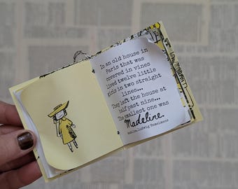 May include: A miniature book with yellow pages featuring a black ink illustration of a character in a yellow coat and hat. The text reads: In an old house in Paris that was covered in vines lived twelve little girls in two straight lines...
