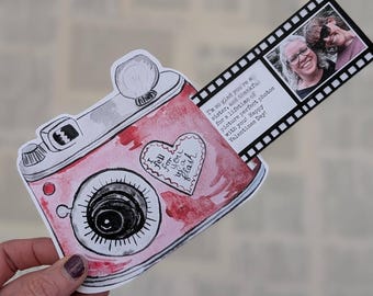 Puede incluir: Una tarjeta en forma de cámara de acuarela rosa y roja con una lente y un flash negros. Una tira de película con dos fotos y el texto "¡Me alegro mucho de que seas mi hermana y agradecida por una vida de fotos perfectas contigo! ¡Feliz día de San Valentín!"