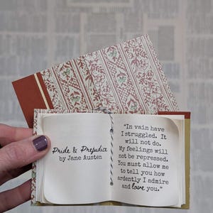 May include: A miniature book titled Pride & Prejudice by Jane Austen with a patterned cover. The open pages display the text: In vain have I struggled. It will not do. My feelings will not be repressed. You must allow me to tell you how ardently I admire and love you.