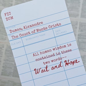 Op de afbeelding: Een witte bibliotheekkaart met blauwe lijnen en rode tekst. De kaart heeft de titel "De graaf van Monte Cristo" van Alexandre Dumas. De tekst "Alle menselijke wijsheid is vervat in deze twee woorden - Wacht en hoop" staat in het rood op de kaart.
