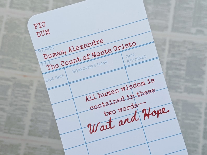 Op de afbeelding: Een witte bibliotheekkaart met blauwe lijnen en rode tekst. De kaart heeft de titel "De graaf van Monte Cristo" van Alexandre Dumas. De tekst "Alle menselijke wijsheid is vervat in deze twee woorden - Wacht en hoop" staat in het rood op de kaart.