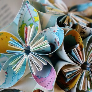 May include: A paper flower ornament made from folded maps. The flower has multiple layers of petals, each with a different section of a map visible. The petals are folded in a way that creates a three-dimensional effect. The flower is a light blue color with yellow, brown, and white accents.