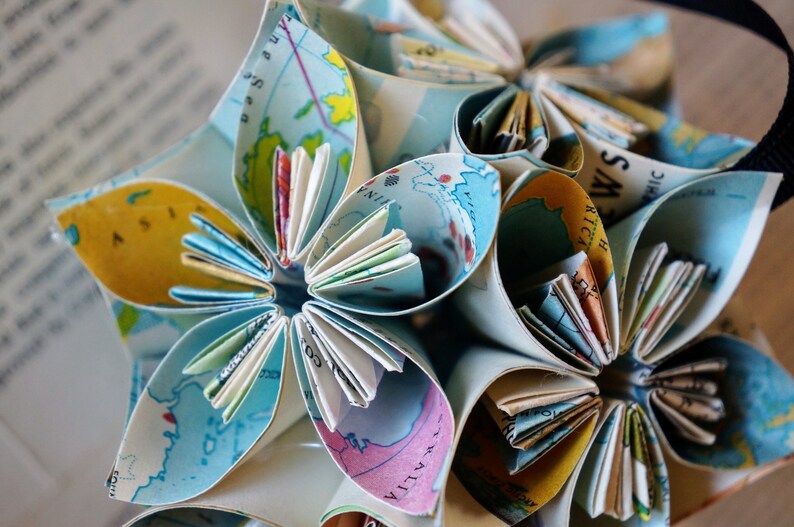 May include: A paper flower ornament made from folded maps. The flower has multiple layers of petals, each with a different section of a map visible. The petals are folded in a way that creates a three-dimensional effect. The flower is a light blue color with yellow, brown, and white accents.