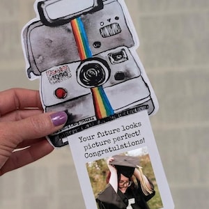May include: A graduation card featuring a watercolor illustration of a vintage Polaroid camera with a rainbow stripe. The card includes a photo of a graduate and the text: "Your future looks picture perfect! Congratulations!"