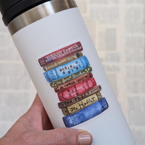 May include: A white Yeti travel mug with a sticker of a stack of books. The sticker features the titles "Harry Potter and the Prisoner of Azkaban", "Anne of Green Gables", "The Secret Garden", "84 Charing Cross Road", "The Thief", and "The Hobbit".