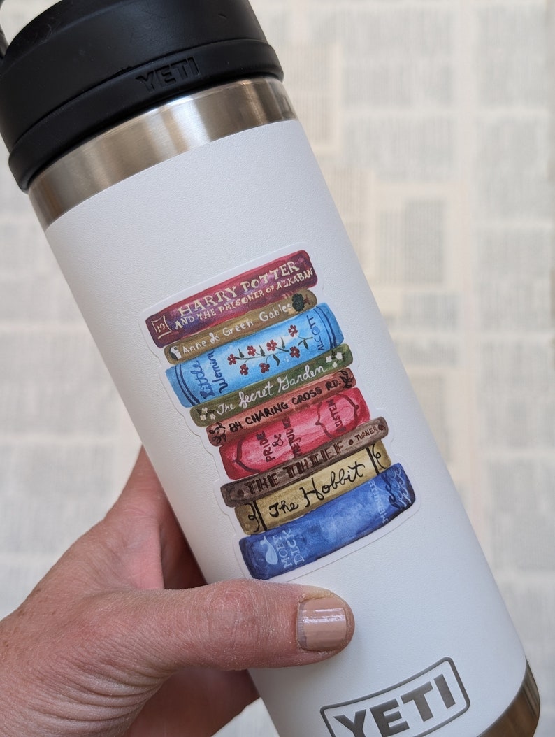May include: A white Yeti travel mug with a sticker of a stack of books. The sticker features the titles "Harry Potter and the Prisoner of Azkaban", "Anne of Green Gables", "The Secret Garden", "84 Charing Cross Road", "The Thief", and "The Hobbit".