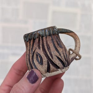 May include: A small, handcrafted ceramic mug featuring a textured, earthy brown surface with deep, carved vertical grooves. The rim is finished with a dark, glossy glaze, and a curved handle is attached to the side with a small piece of twine.