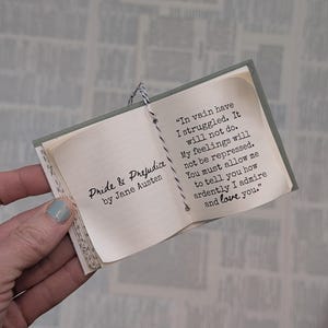 May include: A miniature book titled Pride & Prejudice by Jane Austen, featuring a black and white string binding. The open page displays the text: In vain have I struggled. It will not do. My feelings will not be repressed. You must allow me to tell you how ardently I admire and love you.