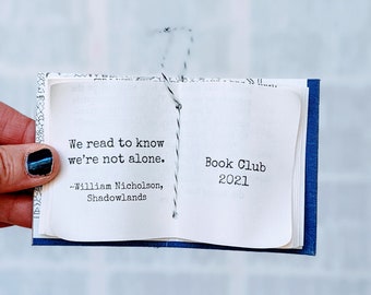 May include: A miniature book with a blue cover featuring a white page printed with the text: We read to know we're not alone. ~William Nicholson, Shadowlands. The right page displays the text: Book Club 2021. A black and white string binds the pages.