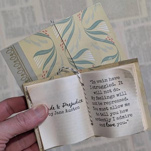 May include: A miniature book titled Pride & Prejudice by Jane Austen with a pale yellow cover featuring a green leaf pattern. The open pages display the text: In vain have I struggled. It will not do. My feelings will not be repressed.