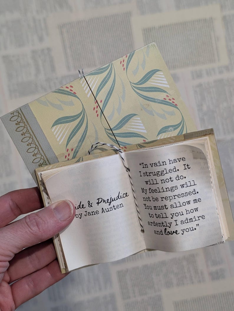 May include: A miniature book titled Pride & Prejudice by Jane Austen with a pale yellow cover featuring a green leaf pattern. The open pages display the text: In vain have I struggled. It will not do. My feelings will not be repressed.